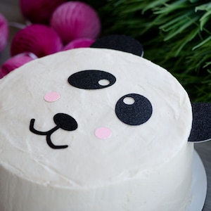 May include: A white cake decorated with black and pink panda bear features. The cake is on a white cake board.