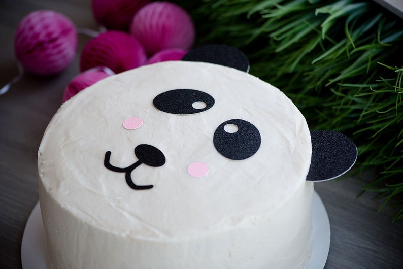 May include: A white cake decorated with black and pink panda bear features. The cake is on a white cake board.