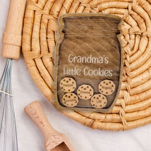 May include: Wooden jar-shaped sign with the text "Grandma's Little Cookies." Inside the jar are wooden cookie cutouts with names: Harper, Emma, Angelo, Dominic, and Lily. The sign is on a woven mat with a whisk and scoop.