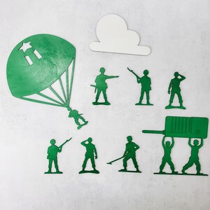 Puede incluir: Un conjunto de soldados de juguete de madera verde en varias poses, incluyendo un soldado con paraca&iacute;das y una nube blanca recortada. Los soldados est&aacute;n dispuestos sobre una superficie blanca.