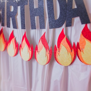May include: A birthday banner with the word "birthday" in black glitter letters and a string of red and yellow flame-shaped decorations.