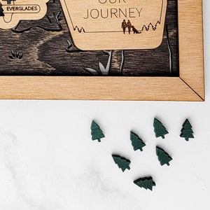 May include: A close-up of a white surface with eight small green wooden pine tree ornaments. The ornaments are scattered on the surface.