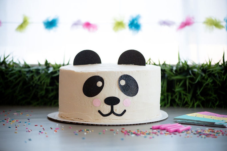 May include: A white cake decorated with black and pink fondant to look like a panda bear. The cake is on a white cake stand and there are colorful sprinkles on the table around it.