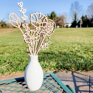 Könnte beinhalten: Eine weiße Keramikvase mit einem Strauß aus lasergeschnittenen Holzblumen. Die Blumen haben die Form von Rosen, Gänseblümchen und Blättern. Die Vase steht auf einem grünen Metalltisch.