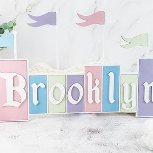 May include: A pastel-colored wooden castle-shaped name sign with the name "Brooklyn" spelled out in white letters. The castle has flags in blue, purple, and green.