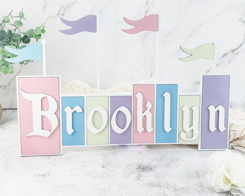 May include: A pastel-colored wooden castle-shaped name sign with the name "Brooklyn" spelled out in white letters. The castle has flags in blue, purple, and green.