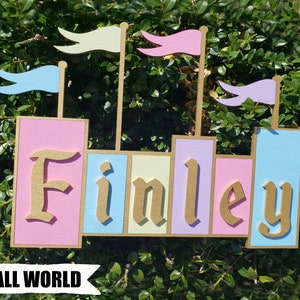 May include: A wooden castle-shaped sign with the name "Finley" in gold lettering. The sign features pastel pink, blue, and yellow blocks, topped with flags. The text "SMALL WORLD" is at the bottom.