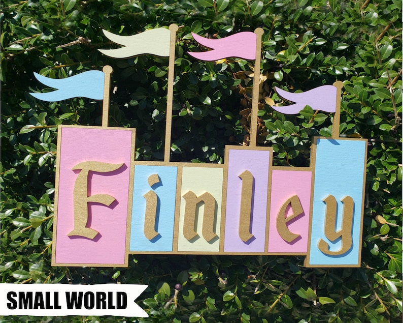 May include: A wooden castle-shaped sign with the name "Finley" in gold lettering. The sign features pastel pink, blue, and yellow blocks, topped with flags. The text "SMALL WORLD" is at the bottom.