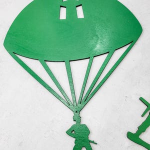 Puede incluir: Un soldado de juguete de madera verde con un paraca&iacute;das. El paraca&iacute;das tiene forma de semic&iacute;rculo con una estrella y dos recortes rectangulares. El soldado est&aacute; de pie con una mano en la cadera y sosteniendo un rifle.