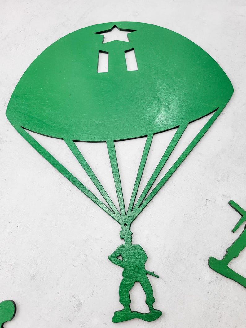 Puede incluir: Un soldado de juguete de madera verde con un paraca&iacute;das. El paraca&iacute;das tiene forma de semic&iacute;rculo con una estrella y dos recortes rectangulares. El soldado est&aacute; de pie con una mano en la cadera y sosteniendo un rifle.