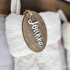 May include: A white fuzzy stocking with a wooden oval tag that says "Joanna" in white script.