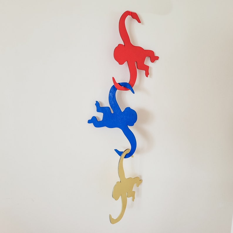 Puede incluir: Tres figuras de monos de madera colgando de una pared blanca. El mono de arriba es rojo, el mono del medio es azul y el mono de abajo es dorado.