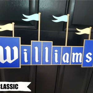 May include: A blue and gold wooden sign in the shape of a castle with the name "Williams" in white lettering. The sign has light blue and yellow flags on top, and the word "CLASSIC" is written at the bottom.