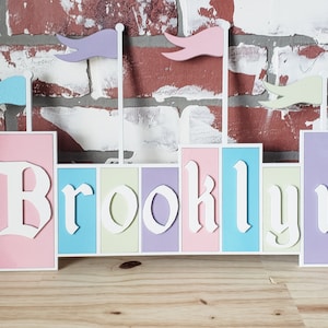 May include: A wooden name sign with the name "Brooklyn" spelled out in white letters. Each letter is on a separate colored block, with the colors pink, blue, cream, purple, and lavender. The sign has four flags on top, with colors purple, pink, blue, and cream.
