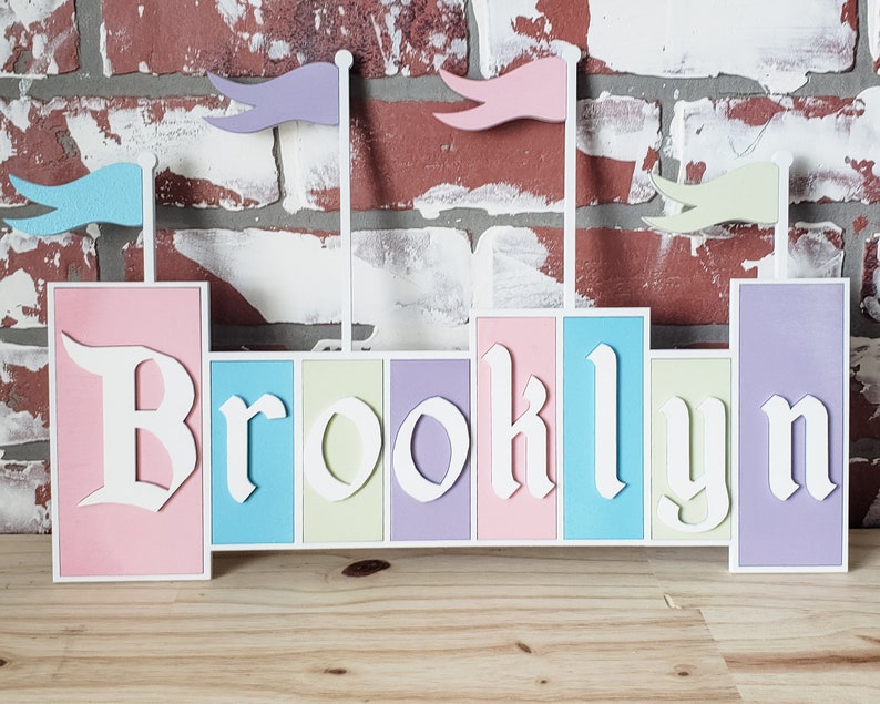 May include: A wooden name sign with the name "Brooklyn" spelled out in white letters. Each letter is on a separate colored block, with the colors pink, blue, cream, purple, and lavender. The sign has four flags on top, with colors purple, pink, blue, and cream.