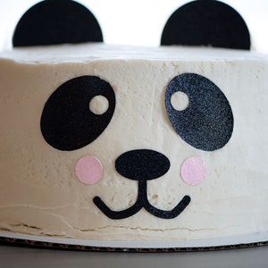May include: A white frosted cake decorated with black and pink paper cutouts to resemble a panda bear face.