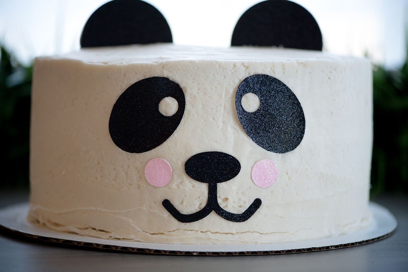 May include: A white frosted cake decorated with black and pink paper cutouts to resemble a panda bear face.