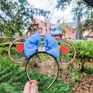 May include: A pair of blue and gold mouse ears with a red rose and a book charm. The ears are decorated with a stained glass design.