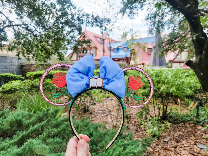 May include: A pair of blue and gold mouse ears with a red rose and a book charm. The ears are decorated with a stained glass design.