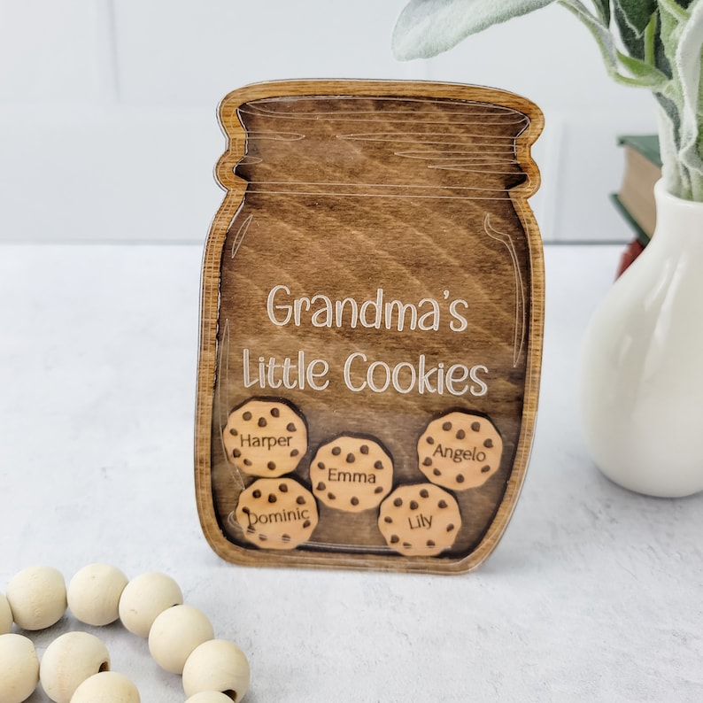 May include: Wooden jar-shaped sign with the words "Grandma's Little Cookies" and the names Harper, Emma, Angelo, Dominic, and Lily on cookie-shaped cutouts. The sign is made of stained wood.