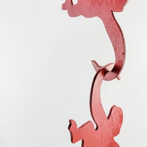 Puede incluir: Una escultura de madera roja de tres figuras, cada una en una pose diferente, conectadas entre s&iacute;. Las figuras son estilizadas y abstractas, asemej&aacute;ndose a formas humanas.