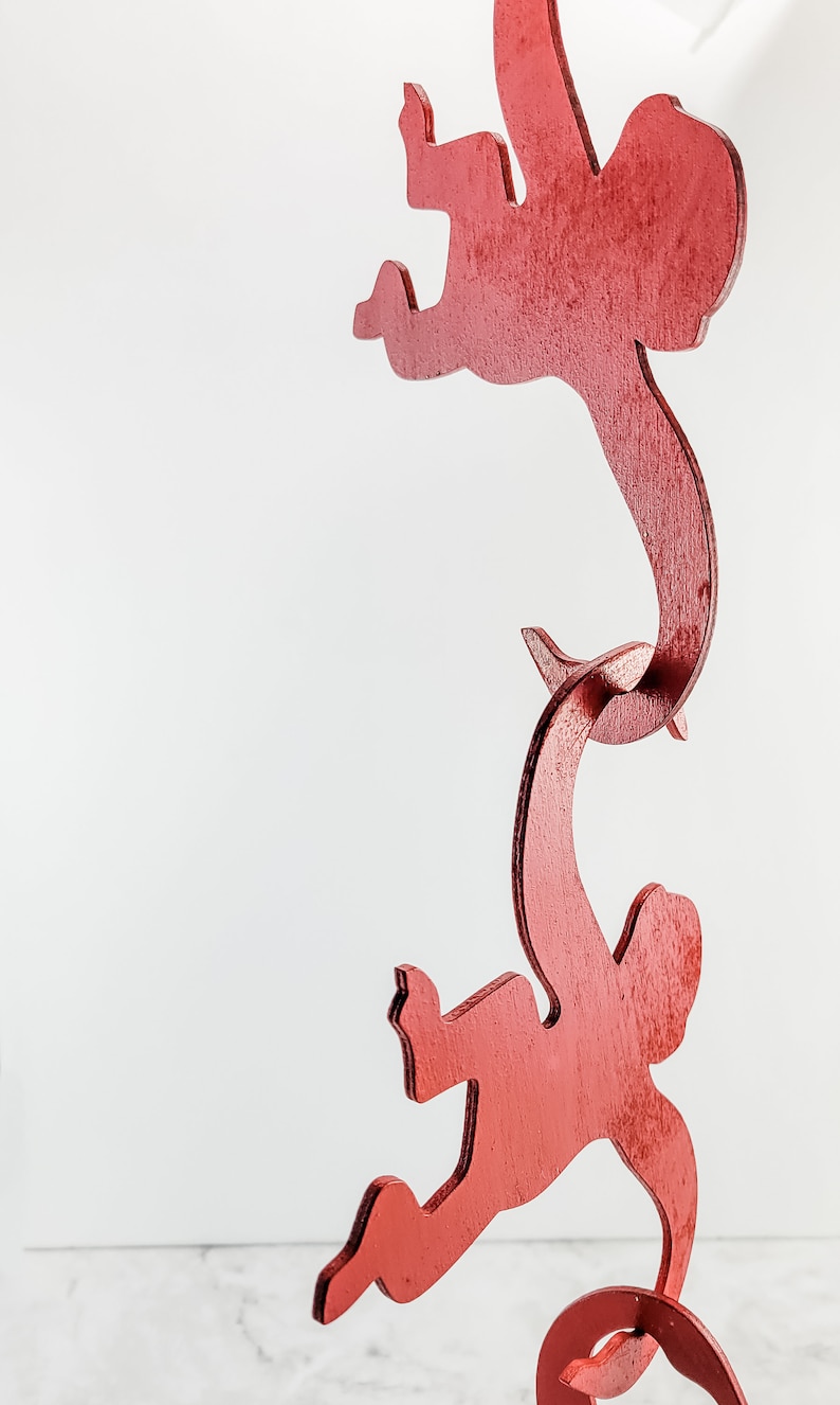 Puede incluir: Una escultura de madera roja de tres figuras, cada una en una pose diferente, conectadas entre s&iacute;. Las figuras son estilizadas y abstractas, asemej&aacute;ndose a formas humanas.