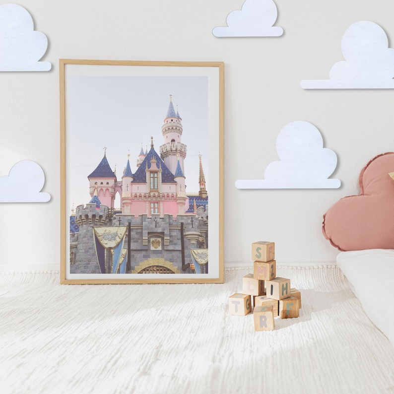 May include: A framed photograph of a pink castle with blue turrets and white clouds on the wall. A stack of wooden blocks with letters on them is on the floor in front of the photo.