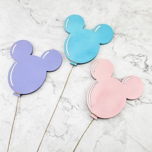 K&ouml;nnte beinhalten: Drei Mickey-Maus-f&ouml;rmige Holzballons in verschiedenen Farben: hellblau, lila und rosa. Die Ballons sind an Schn&uuml;ren befestigt.