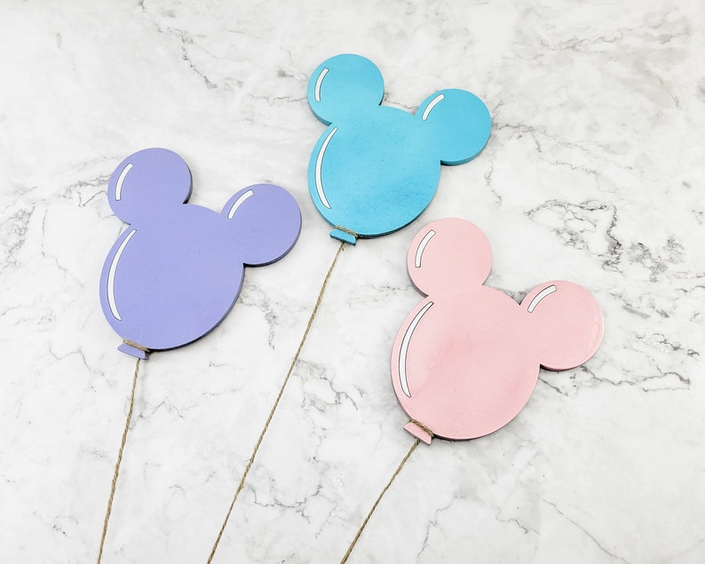 K&ouml;nnte beinhalten: Drei Mickey-Maus-f&ouml;rmige Holzballons in verschiedenen Farben: hellblau, lila und rosa. Die Ballons sind an Schn&uuml;ren befestigt.