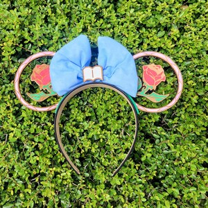 May include: A pair of blue and pink mouse ears with a gold and green rose design. The ears are decorated with a blue bow and a book charm.