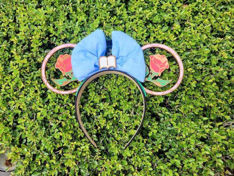 May include: A pair of blue and pink mouse ears with a gold and green rose design. The ears are decorated with a blue bow and a book charm.