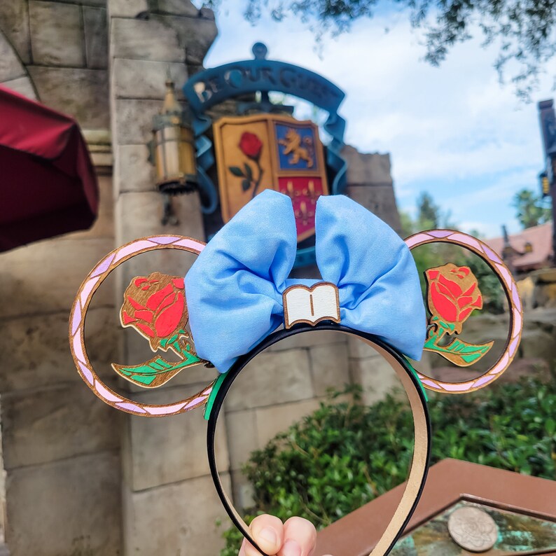 May include: A pair of light blue fabric Minnie Mouse ears with a white book charm. The ears are decorated with a pink and purple wooden frame with a red rose design.