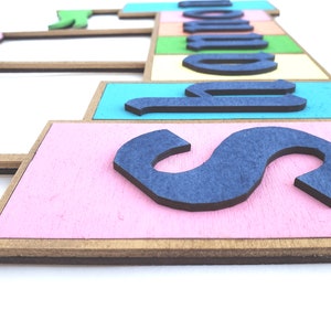 May include: A wooden sign with the word "SCHOOL" spelled out in blue letters. The letters are on colorful, pastel-colored rectangular blocks. The sign has a pink, blue, green, and yellow color scheme.