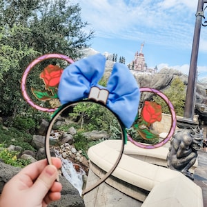 May include: A pair of blue and pink Minnie Mouse ears with a blue bow and a rose design. The ears are in front of a castle and a waterfall.