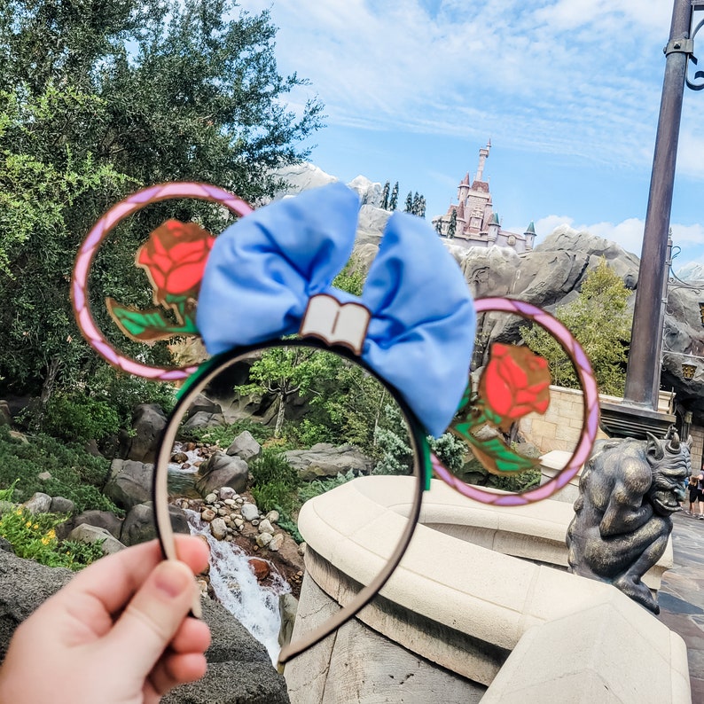 May include: A pair of blue and pink Minnie Mouse ears with a blue bow and a rose design. The ears are in front of a castle and a waterfall.
