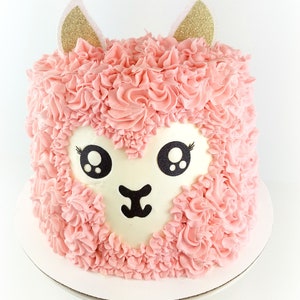 May include: A pink llama cake with gold glitter ears. The cake is decorated with pink frosting and has a white face with black eyes and a nose.
