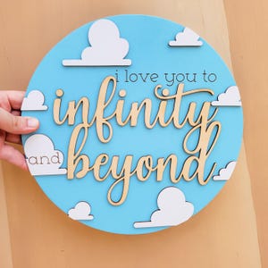 May include: A round, light blue wooden sign with white cloud cutouts. The sign features the phrase "i love you to infinity and beyond" in a cursive font. The sign is a decorative item.