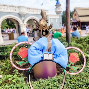 May include: A pair of light blue Minnie Mouse ears with a gold-coloured headband. The ears are decorated with a large blue bow and a gold-coloured book charm. Each ear features a gold-coloured frame with a red rose inside.