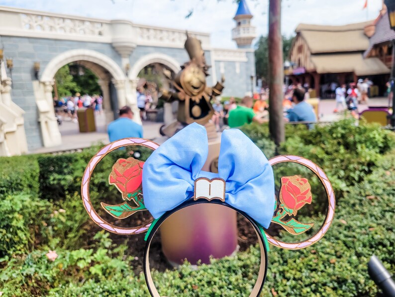 May include: A pair of light blue Minnie Mouse ears with a gold-coloured headband. The ears are decorated with a large blue bow and a gold-coloured book charm. Each ear features a gold-coloured frame with a red rose inside.