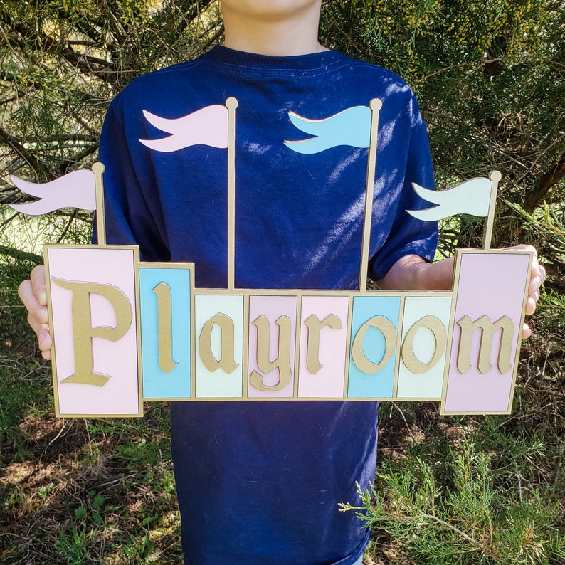 May include: A wooden sign with the word "Playroom" in gold lettering. The sign is designed to resemble a castle, with pink, blue, and mint green panels. Three small flags are attached to the top of the sign.