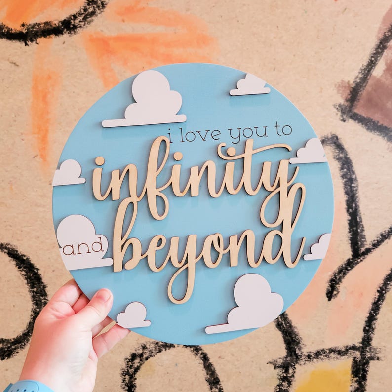 May include: A round, light blue wooden sign with the phrase "I love you to infinity and beyond" in cursive script. White cloud cutouts are placed around the edge of the sign. The sign is held by a hand.