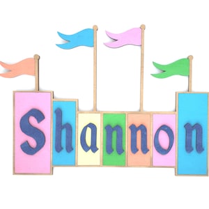May include: A colorful wooden castle-themed name sign with the name "Shannon" in blue letters. The sign features pink, blue, yellow, and green blocks, with flags on top. A decorative item for a child's room.