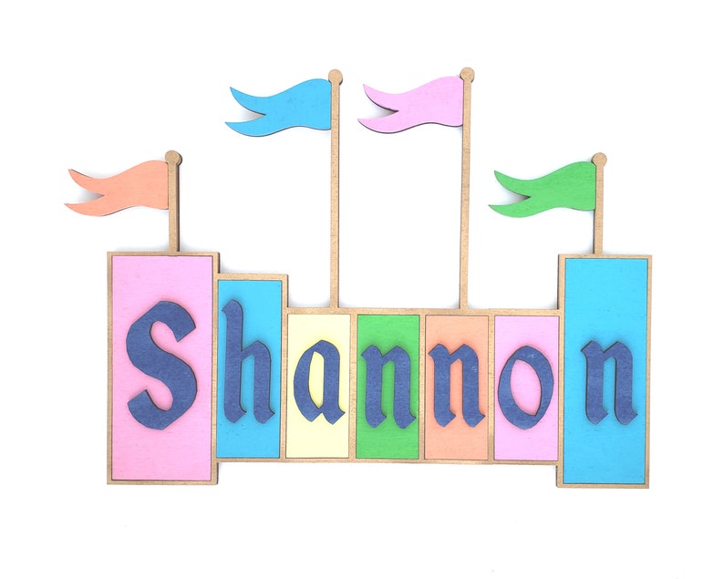 May include: A colorful wooden castle-themed name sign with the name "Shannon" in blue letters. The sign features pink, blue, yellow, and green blocks, with flags on top. A decorative item for a child's room.