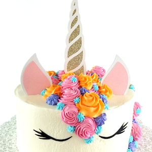 May include: A white cake decorated with pink, orange, and blue frosting in the shape of a unicorn. The cake has a gold glitter horn and pink ears.