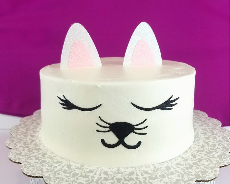May include: White cake decorated with a cat face. The cat has black eyes, a black nose, and black whiskers. The cake has pink ears on top.
