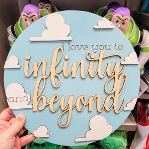 May include: A round, light blue wooden sign with the phrase "I love you to infinity and beyond" in cursive lettering. White cloud cutouts are placed around the edge of the sign. The sign is a decorative item.