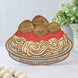 Könnte beinhalten: Holzausschnitt eines Tellers mit Spaghetti und Fleischbällchen, auf denen die Namen "Caeden", "Norah", "Caleb" und "Henry" stehen. Der Text "Grandma's Little Meatballs" steht am unteren Rand des Tellers.