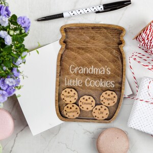 May include: A wooden jar-shaped sign with the text "Grandma's Little Cookies." The sign has a clear front and contains wooden cookie cutouts with names. A pen, flowers, and a gift are in the background.