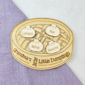 Könnte beinhalten: Holzzeichen mit einer Zeichnung eines Dampfkorbs und vier Holzklößen mit den Namen Leilani, Kai, Mina und Naomi. Das Schild lautet "Grandma's Little Dumplings".