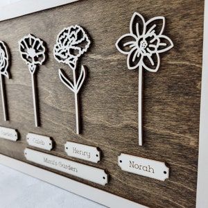 Puede incluir: Una placa de madera con cinco flores recortadas en blanco y nombres debajo de cada flor. Los nombres son "Garden", "Caleb", "Henry", "Mimi's Garden" y "Norah".